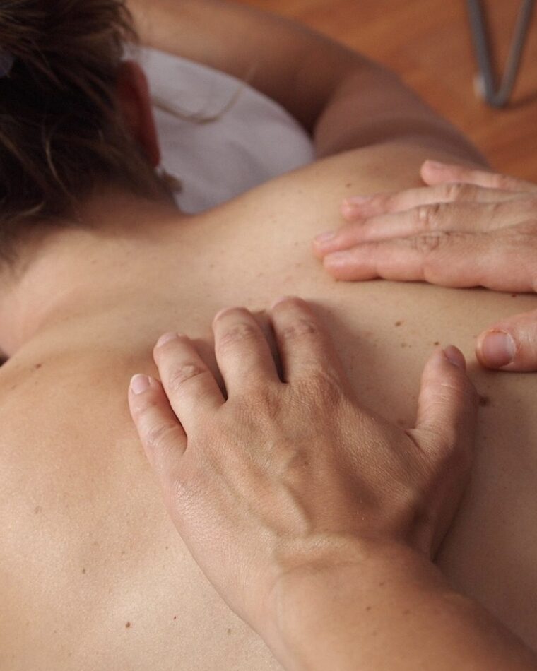A therapist working with both hands across a client's upper back and shoulder during a therapeutic massage session.